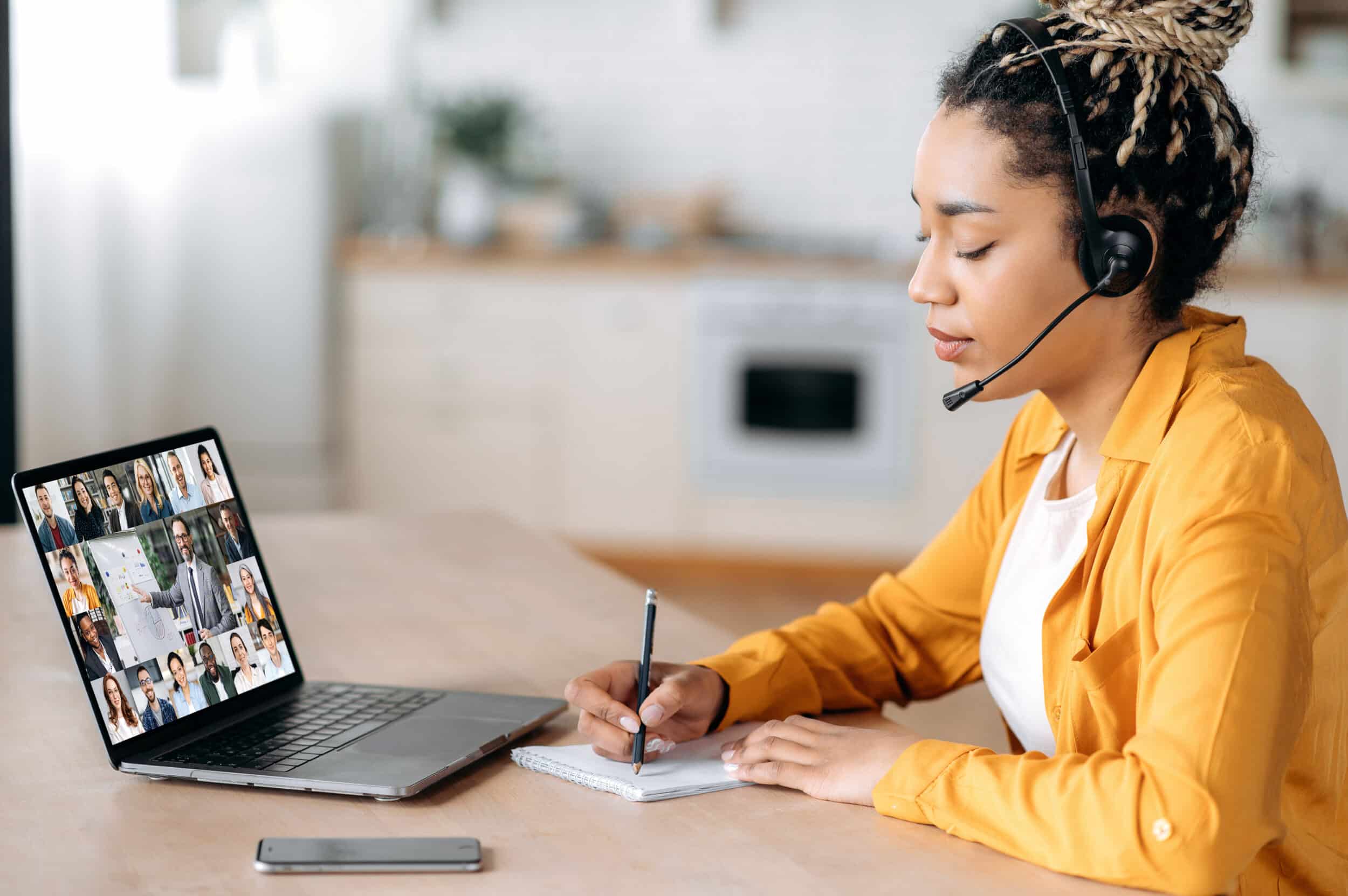 Online webinar, business meeting. Side view of african american woman, communicates with business colleagues by video call uses laptop, takes notes, multiracial coworkers on laptop screen, brainstorm